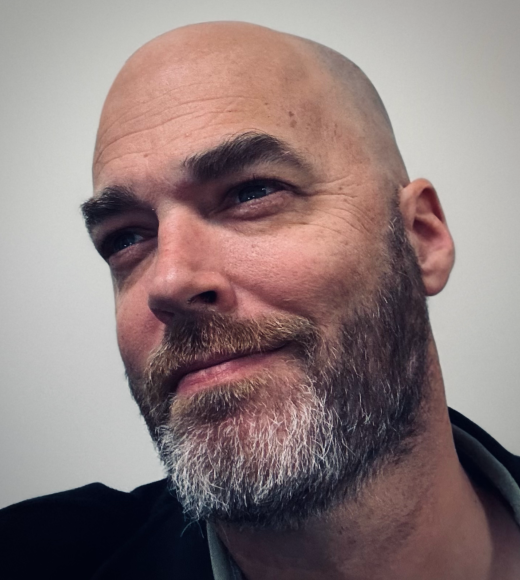 A man with a bald head and a short gray beard, looking to the side with a slight smile.