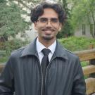 Man smiling in glasses and dark jacket by wooden railing outdoors (photograph)